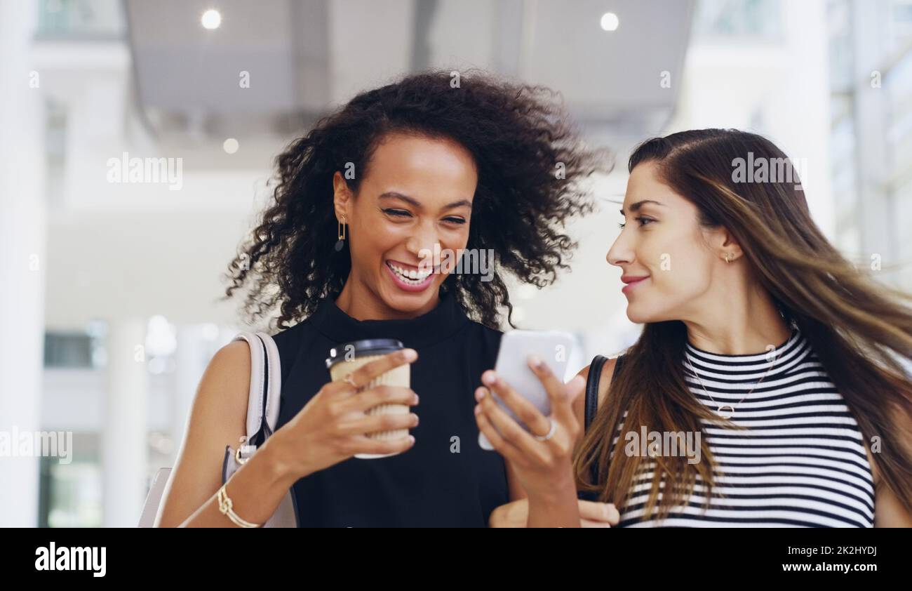 Sono il meglio dei colleghi. Scatto corto di due giovani donne d'affari che chiacchierano e usano uno smartphone mentre camminano in un ufficio durante una pausa caffè. Foto Stock