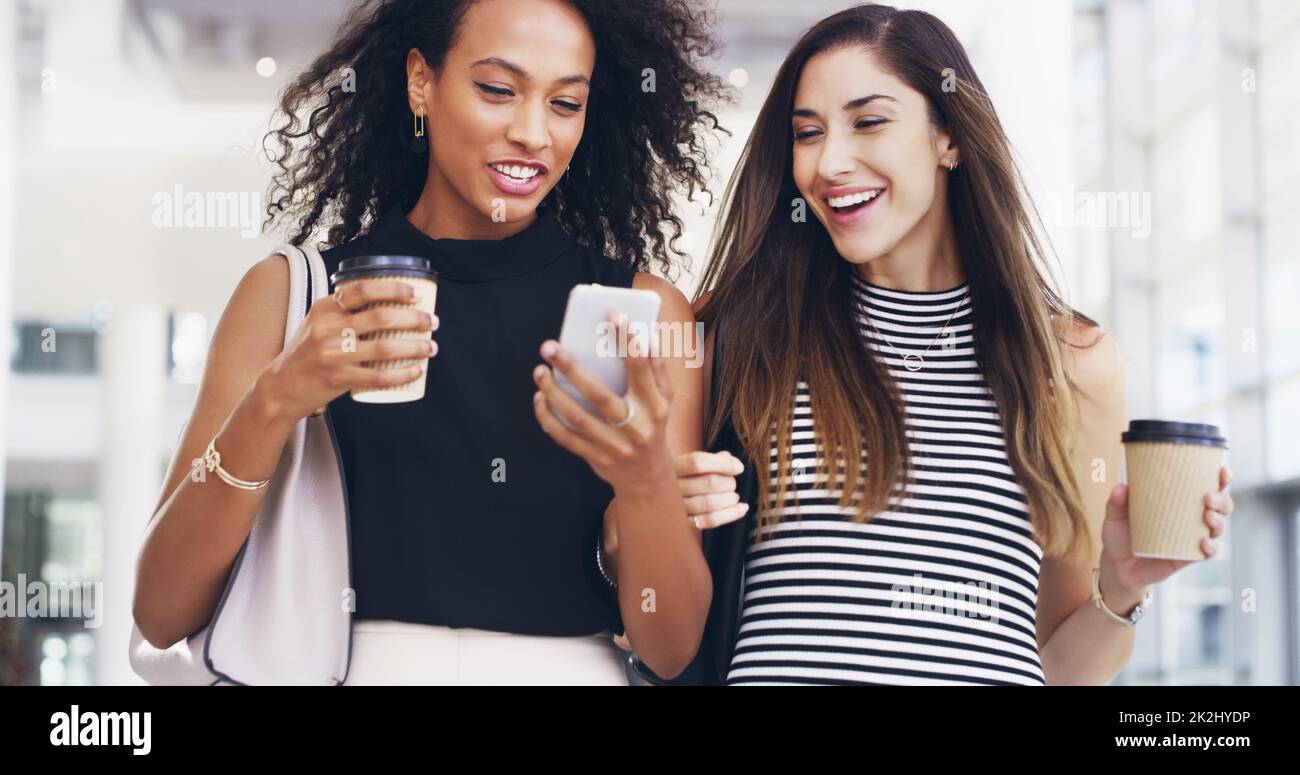 Condividono tutto. Scatto corto di due giovani donne d'affari che chiacchierano e usano uno smartphone mentre camminano in un ufficio durante una pausa caffè. Foto Stock