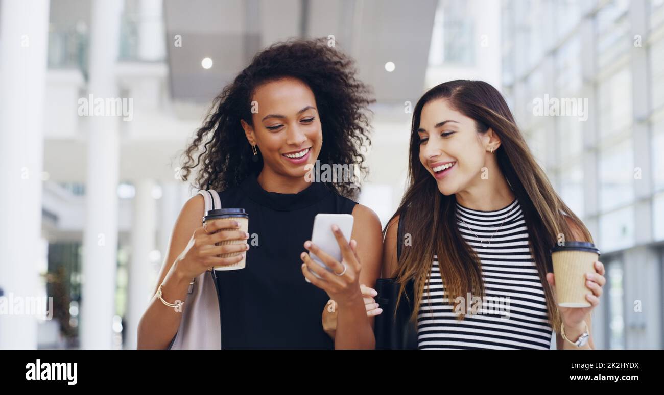 Fantastico. Scatto corto di due giovani donne d'affari che chiacchierano e usano uno smartphone mentre camminano in un ufficio durante una pausa caffè. Foto Stock