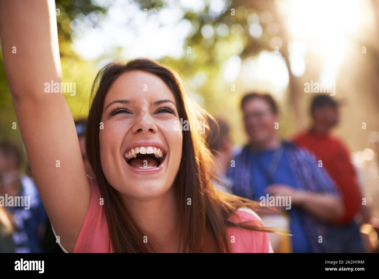 È una orgogliosa sostenitrice. Una bella giovane donna sorridente e godendo di musica in un festival con braccio sollevato in aria e folla in background. Foto Stock