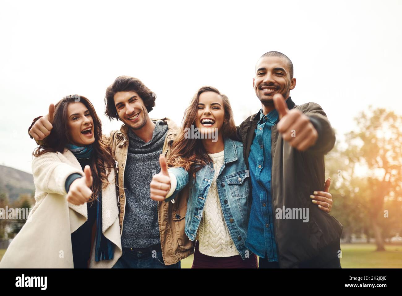 Che giornata fantastica da trascorrere con gli amici. Ritratto di un gruppo di giovani amici allegri accoccolati insieme, mostrando i pollici all'esterno durante il giorno. Foto Stock