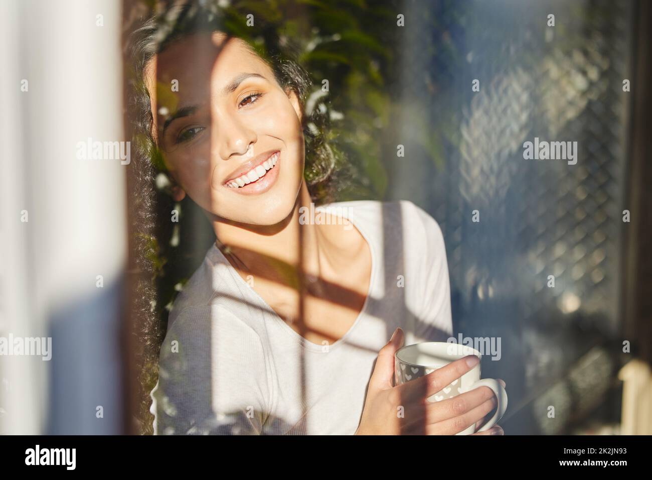 Adoro la sensazione di sole mattutino. Scatto di una giovane donna in piedi e godere di una tazza di caffè guardando fuori dalla sua finestra. Foto Stock