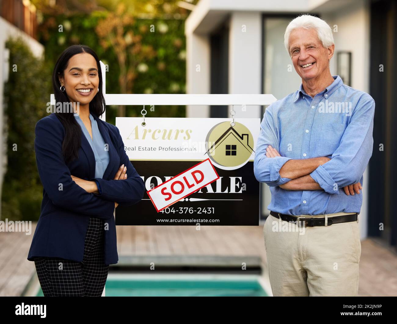 Ritratto corto di un agente immobiliare giovane attraente e il suo cliente maschio in piedi con le braccia incrociate accanto a un segno venduto al di fuori della sua casa appena acquistata Foto Stock