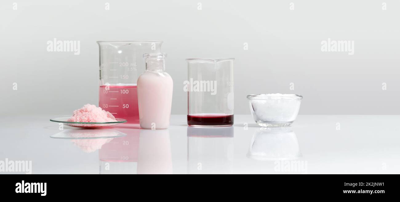Crema per la cura della bellezza, prodotti chimici in scaglie rosa, permanganato di potassio liquido e cera di esteri di cetile su tavolo da laboratorio bianco. Foto Stock