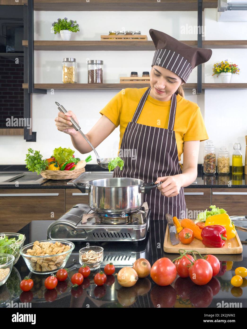 Giovane casalinga asiatica vestita con un grembiule e un cappellino per capelli, preparando la zuppa di verdure in una cucina moderna. Foto Stock