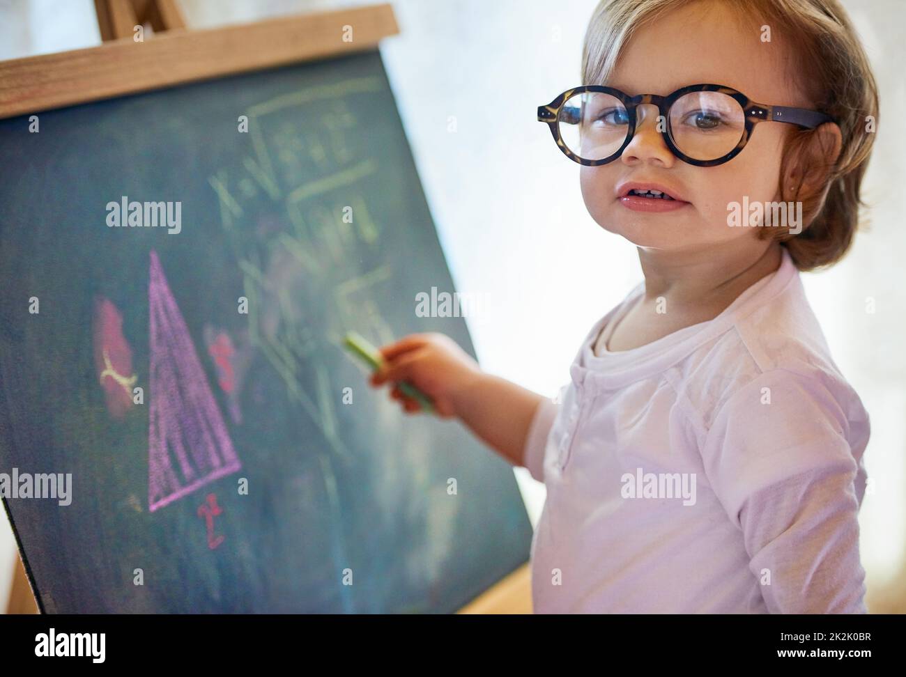 Fare la matematica assomiglia ai bambini giocare. Ritratto di una bambina adorabile con grandi occhiali che risolve un problema di matematica su una lavagna a casa. Foto Stock