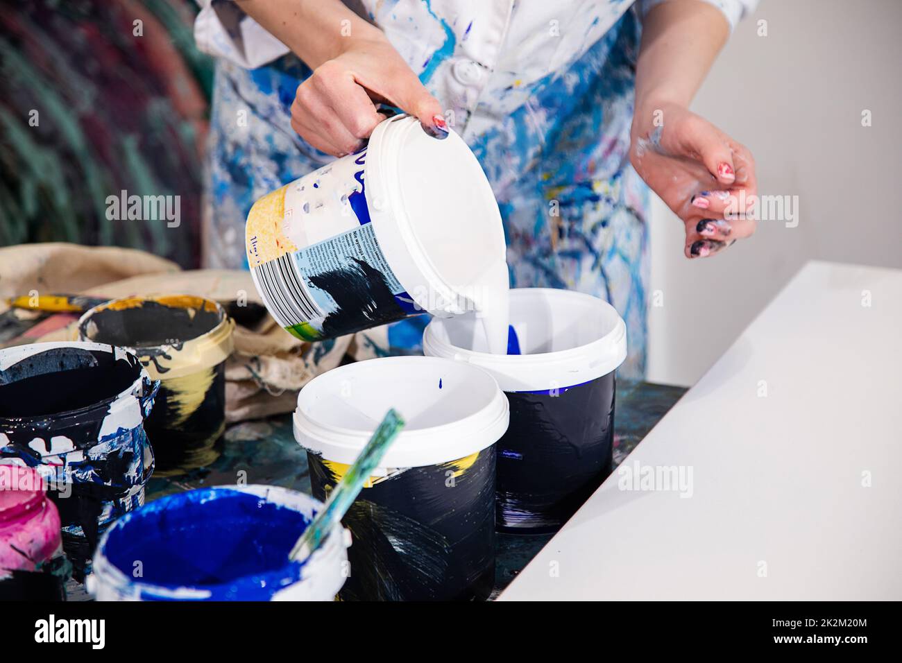 Foto ritagliata delle mani della donna in vestito ricoperto di macchie versando vernice acrilica bianca dal secchio in un altro secchio. Foto Stock
