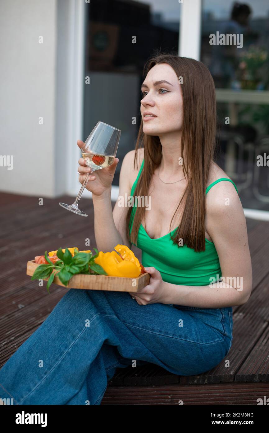 Ritratto di giovane donna pensierosa splendida seduta su un gradino di veranda in legno, tenendo un bicchiere di vino, verdure tagliate. Foto Stock