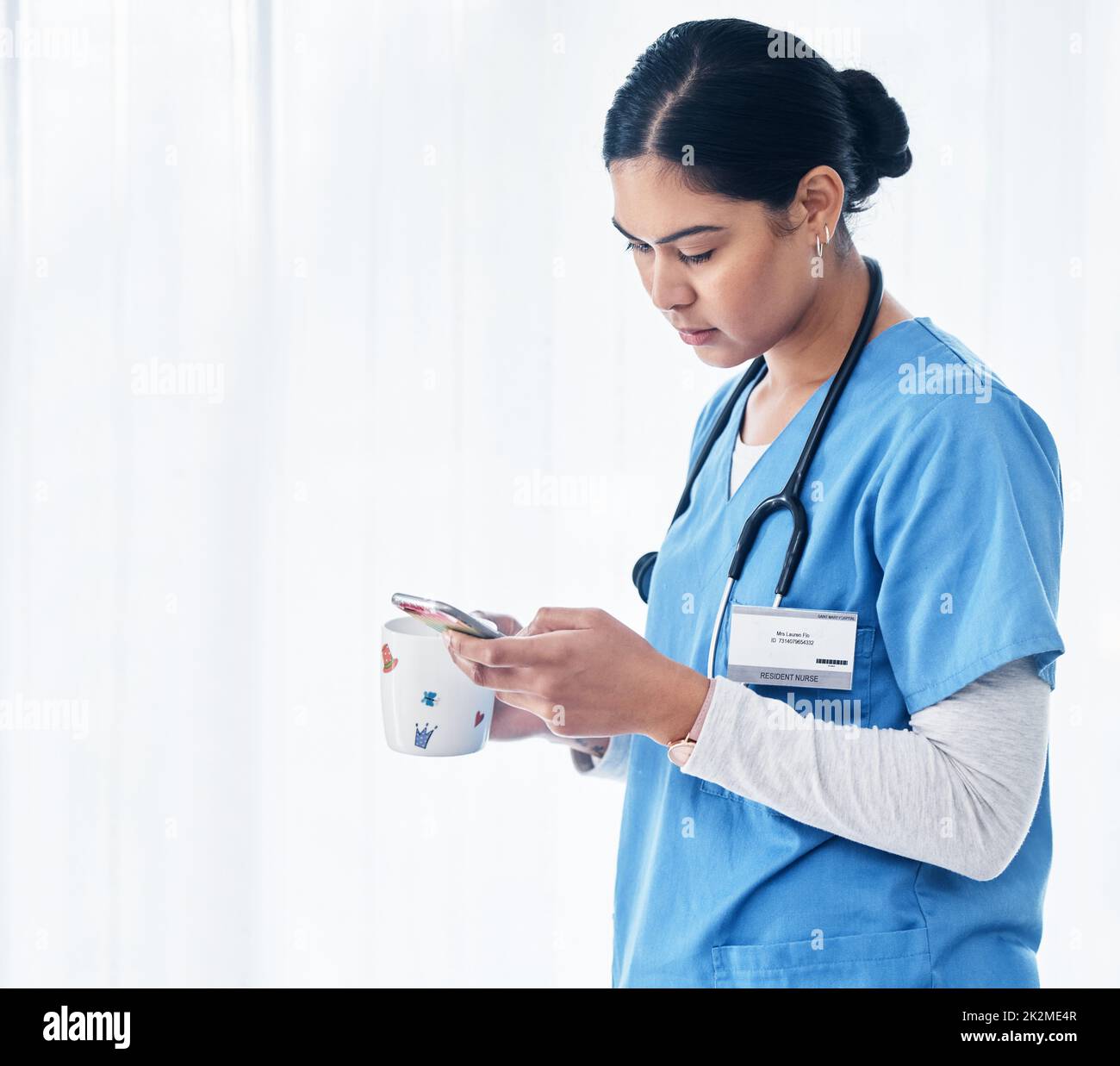 Chatta ora in pausa. Shot di un'infermiera femminile che tiene una tazza di caffè mentre usa il suo cellulare. Foto Stock