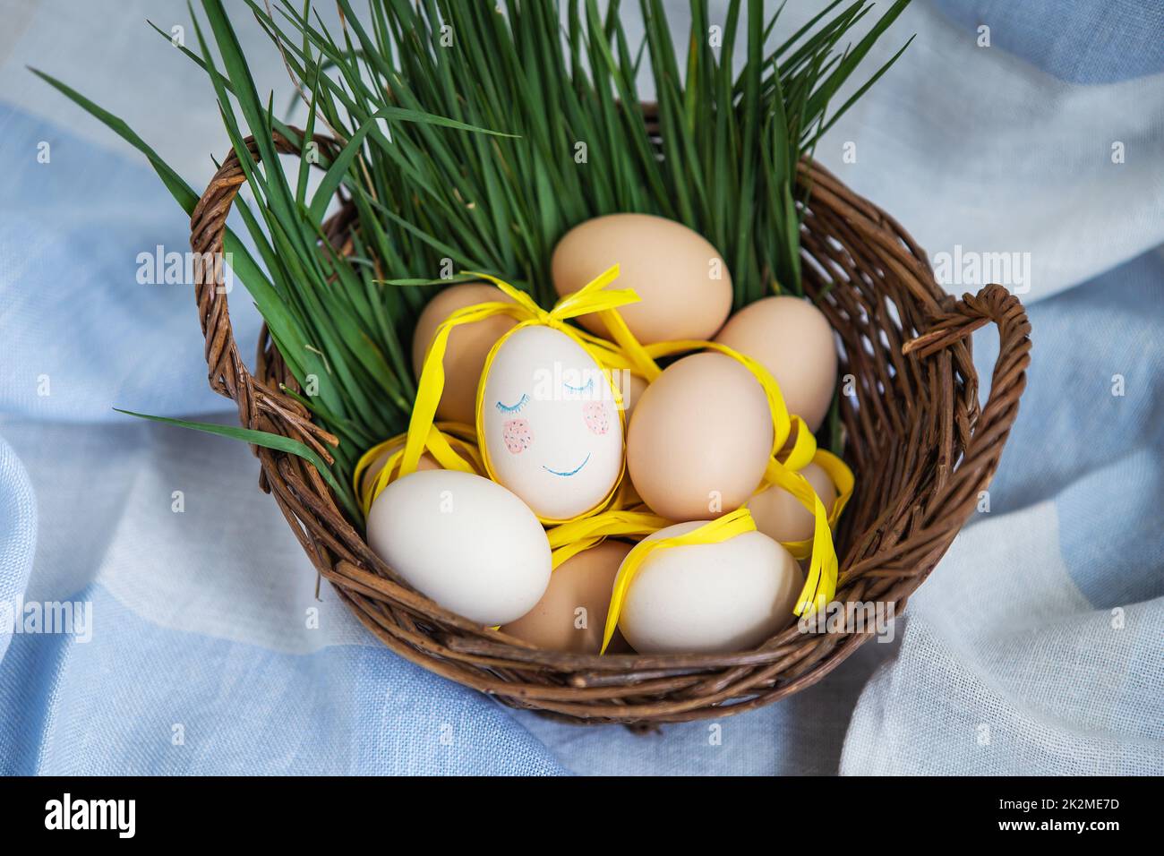 Uova di Pasqua dipinte, un uovo con un viso carino, le uova giacciono in un cesto di legno insieme all'erba verde. Cartolina di Pasqua. Foto Stock