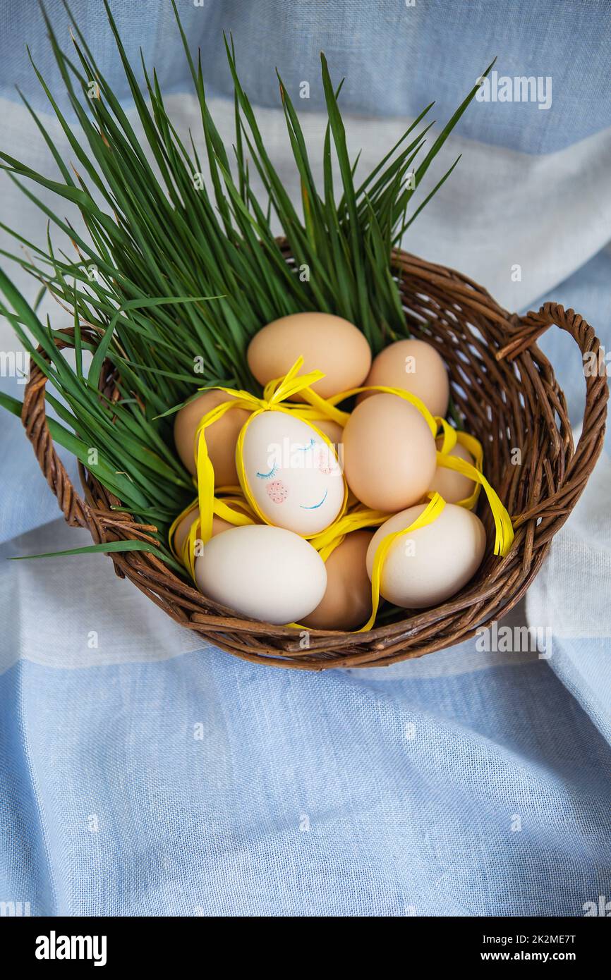 Uova di Pasqua dipinte, un uovo con un viso carino, le uova giacciono in un cesto di legno insieme all'erba verde. Cartolina di Pasqua. Foto Stock