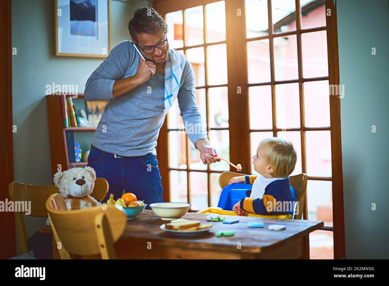 Daddy sa come operare in multitasking. Scatto corto di un singolo padre su una chiamata mentre alimenta suo figlio a casa. Foto Stock