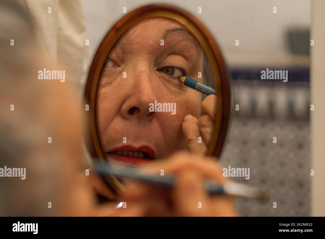una donna più anziana dipinge la sua linea dell'occhio davanti ad uno specchio Foto Stock