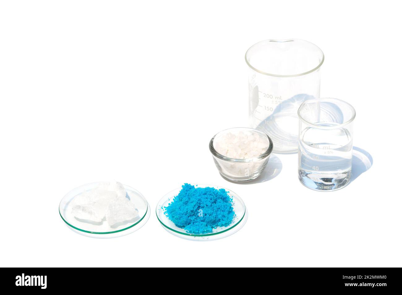 Cristalli di allume e solfato di rame(II) in vetro da orologio chimico su tavolo da laboratorio bianco. Il sale di scaglie in un contenitore di vetro e l'alcool in un becher sono stati posti sullo sfondo. Foto Stock