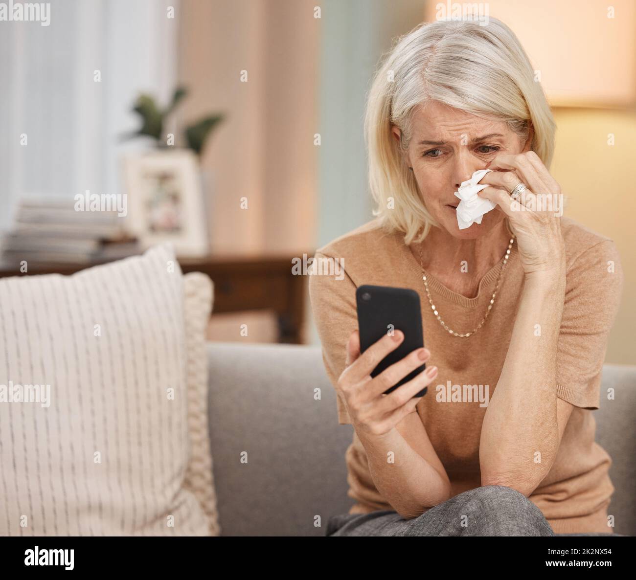 2020, l'anno segnato da trauma. Scatto di una donna matura utilizzando uno smartphone e piangendo sul divano a casa. Foto Stock