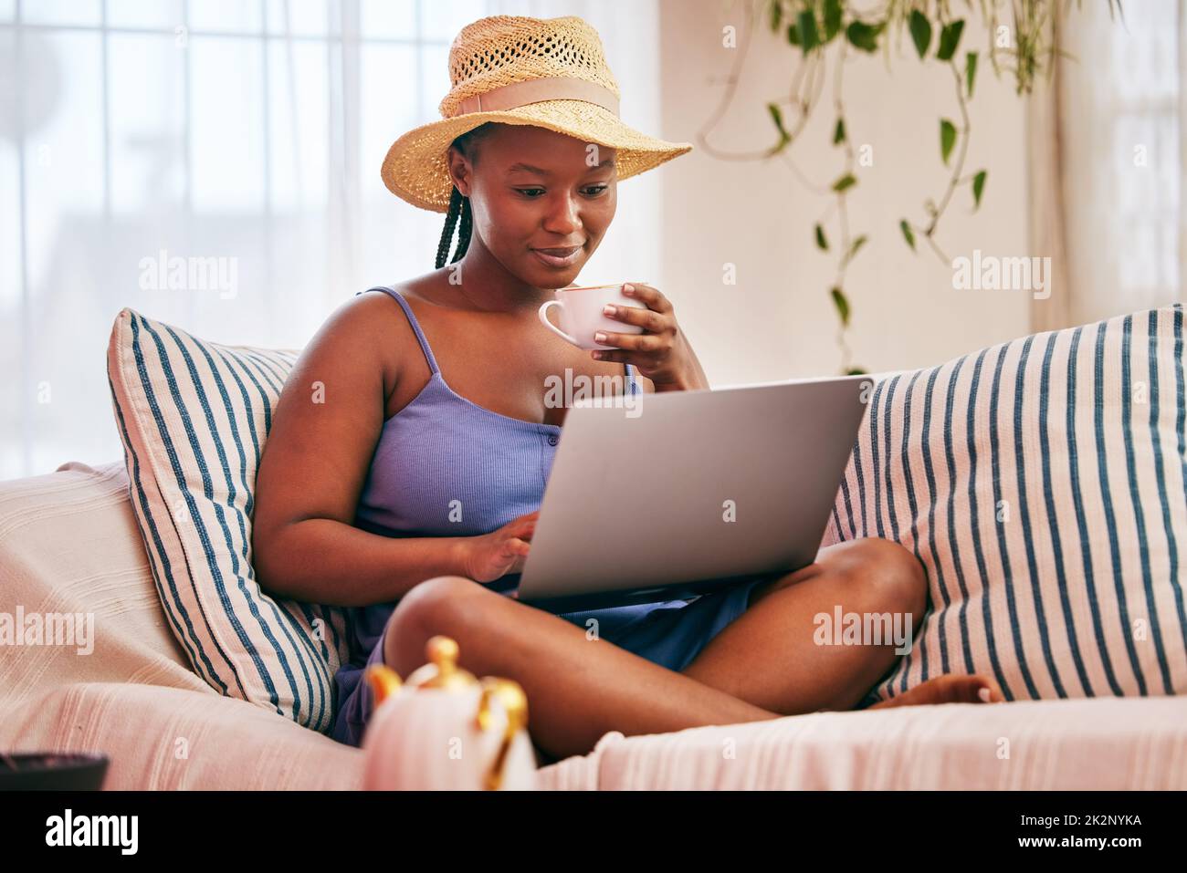 Theres nessun posto come casa. Scatto di una giovane donna che indossa un cappello da sole e usa il suo laptop mentre fa una pausa caffè sul divano. Foto Stock
