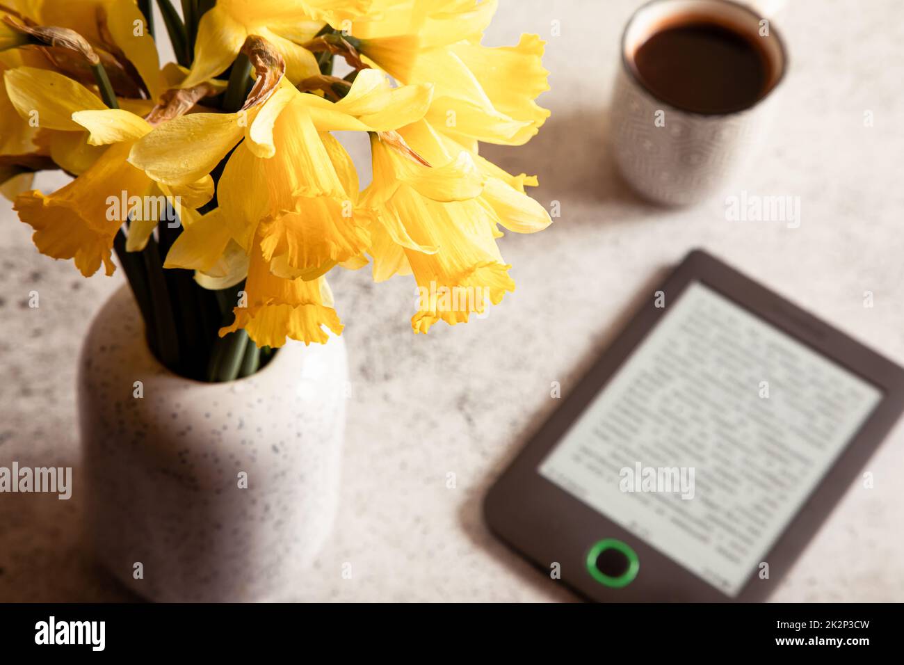 Moderno lettore di ebook, fiori di daffodils sullo sfondo. Vista dall'alto, composizione minima in stile di posa piatta. Scrivania donna, blogger moda, tecnologia di bellezza Foto Stock
