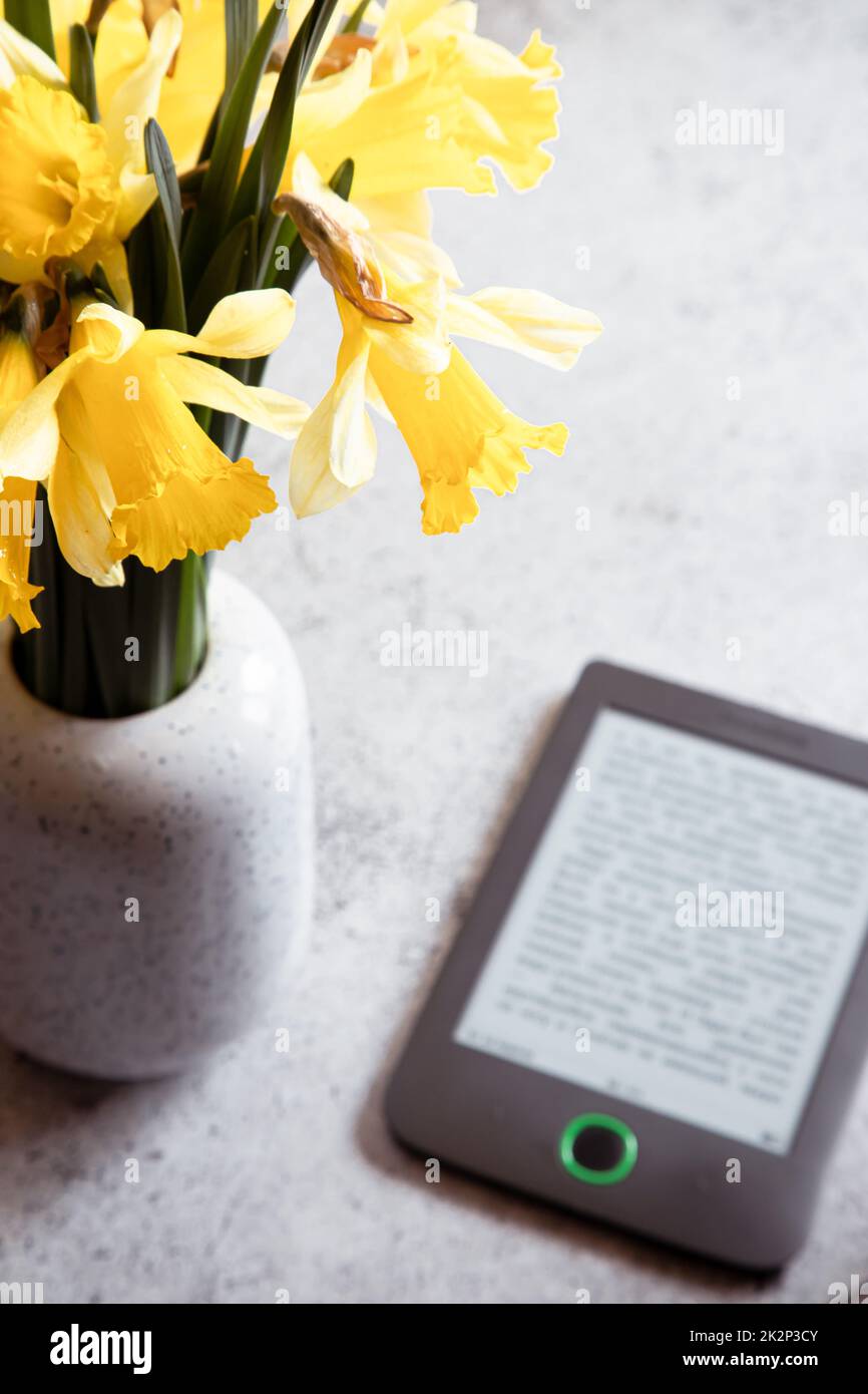 Moderno lettore di ebook, fiori di daffodils sullo sfondo. Vista dall'alto, composizione minima in stile di posa piatta. Scrivania donna, blogger moda, tecnologia di bellezza Foto Stock