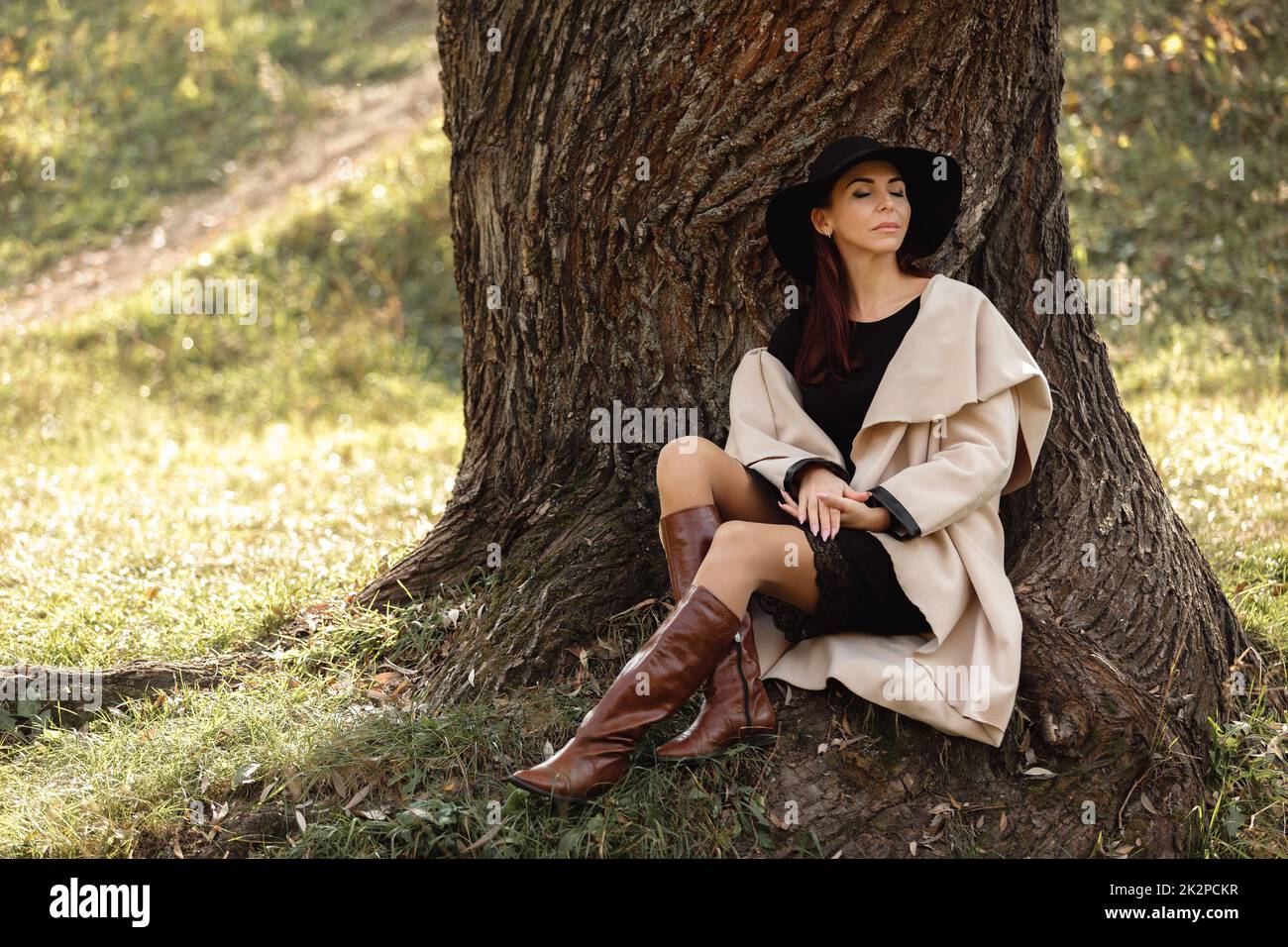 La donna in un cappotto beige e cappello nero seduto sotto agli alberi Foto Stock