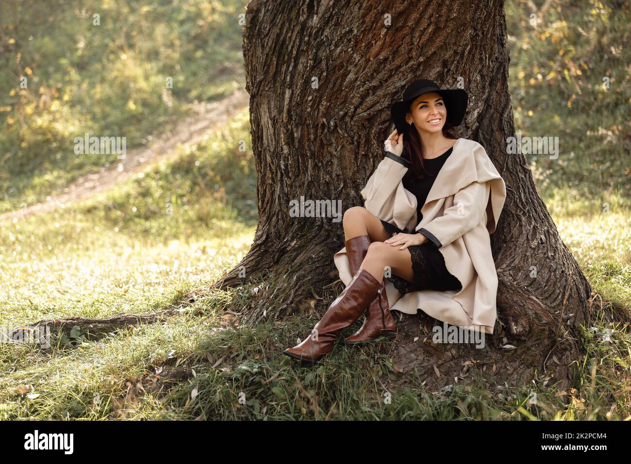 La donna in un cappotto beige e cappello nero seduto sotto agli alberi Foto Stock