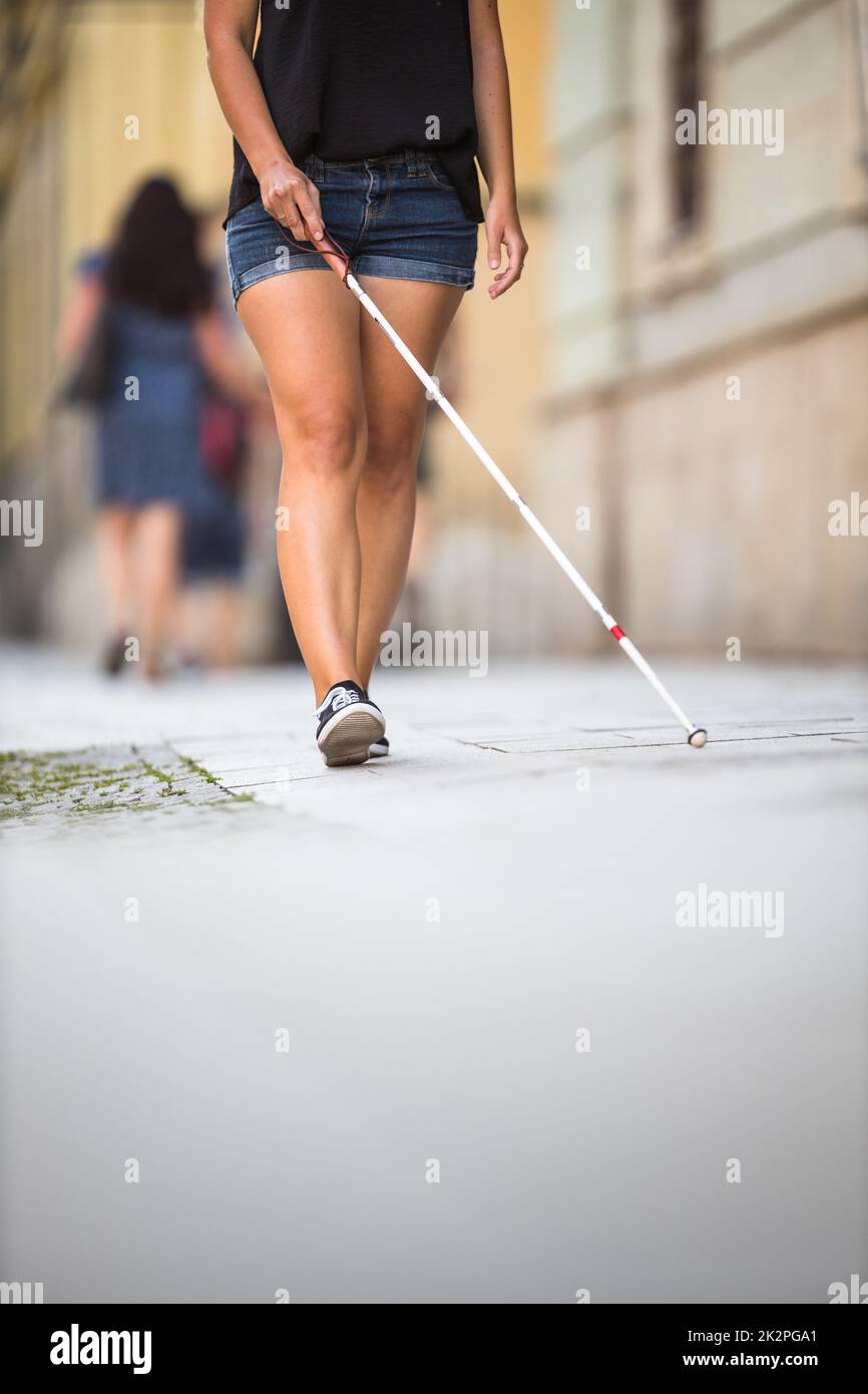 Donna cieca che cammina per le strade della città, utilizzando la sua canna bianca per navigare meglio lo spazio urbano e per raggiungere la sua destinazione in modo sicuro Foto Stock