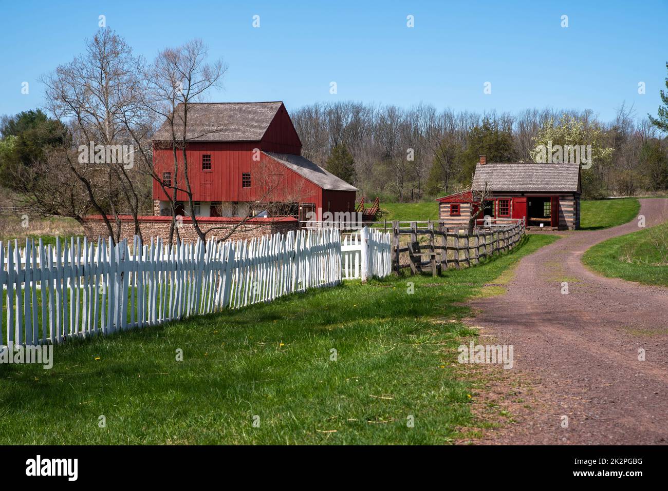 Strada sterrata con recinzione da picket e un fienile coloniale americano rosso e capanna di tronchi Foto Stock