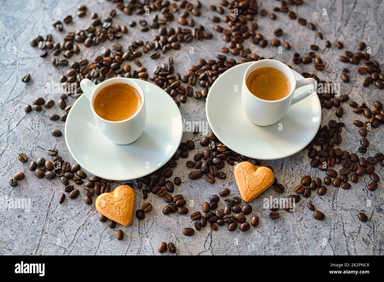 Due tazze di caffè espresso con piattini su un tavolo in pietra con biscotti al cuore e chicchi di caffè torrefatti. Foto Stock