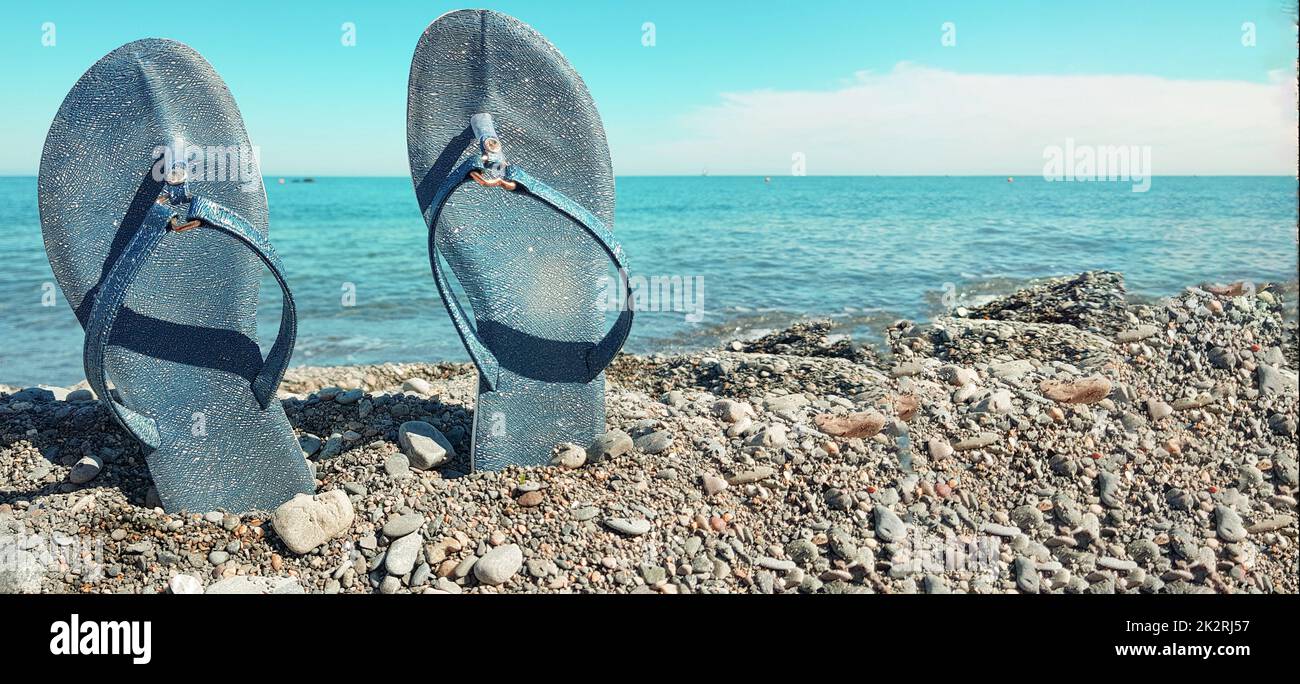 Scarpe da spiaggia blu da donna, in piedi nei ciottoli sullo sfondo del mare e del cielo blu. Primo piano, posto per il testo, concetto di una vacanza al mare, banner Foto Stock