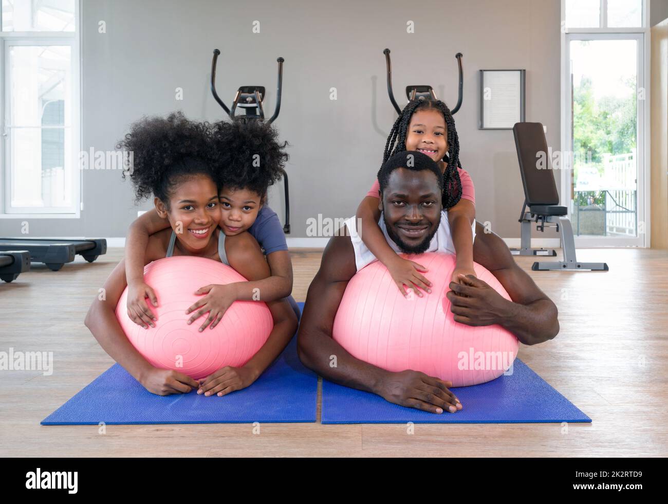 Il giovane capello nero riccio e sua moglie si rilassano su una palla di yoga mentre entrambe le figlie sono sulla schiena. Famiglia felice godere insieme vacanza nel centro fitness. Foto Stock