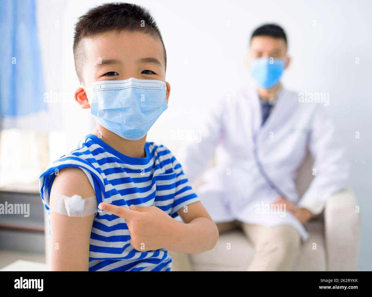 Bambino felice che indossa maschera medica e spalla con bendaggio adesivo sul braccio dopo la vaccinazione Foto Stock