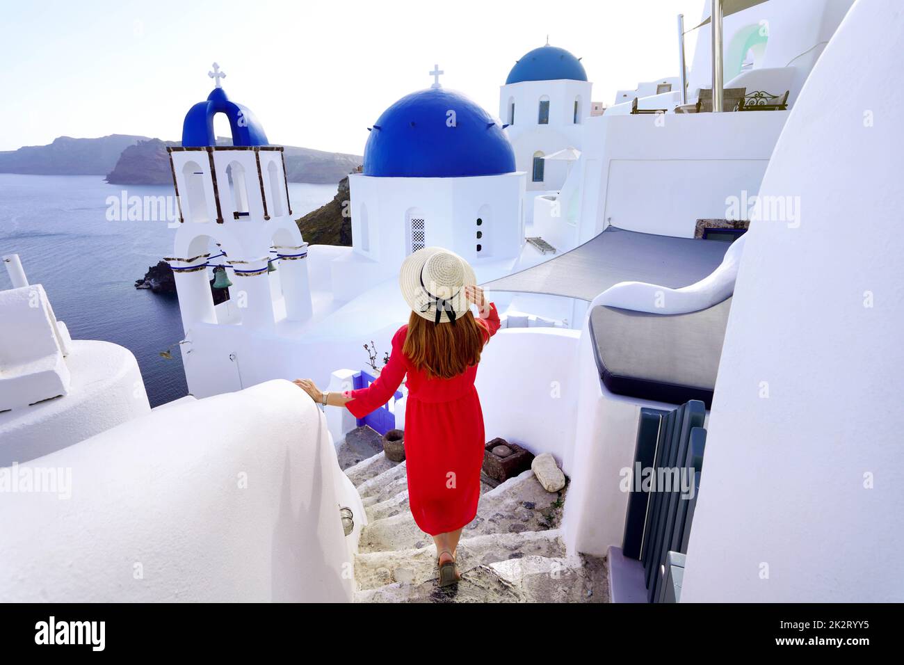 Turismo a Santorini. Giovane donna alla moda con vestito rosso e cappello scende lungo i gradini nel villaggio di Oia, sull'isola di Santorini. Turista di viaggio femminile per le sue vacanze in Grecia. Foto Stock
