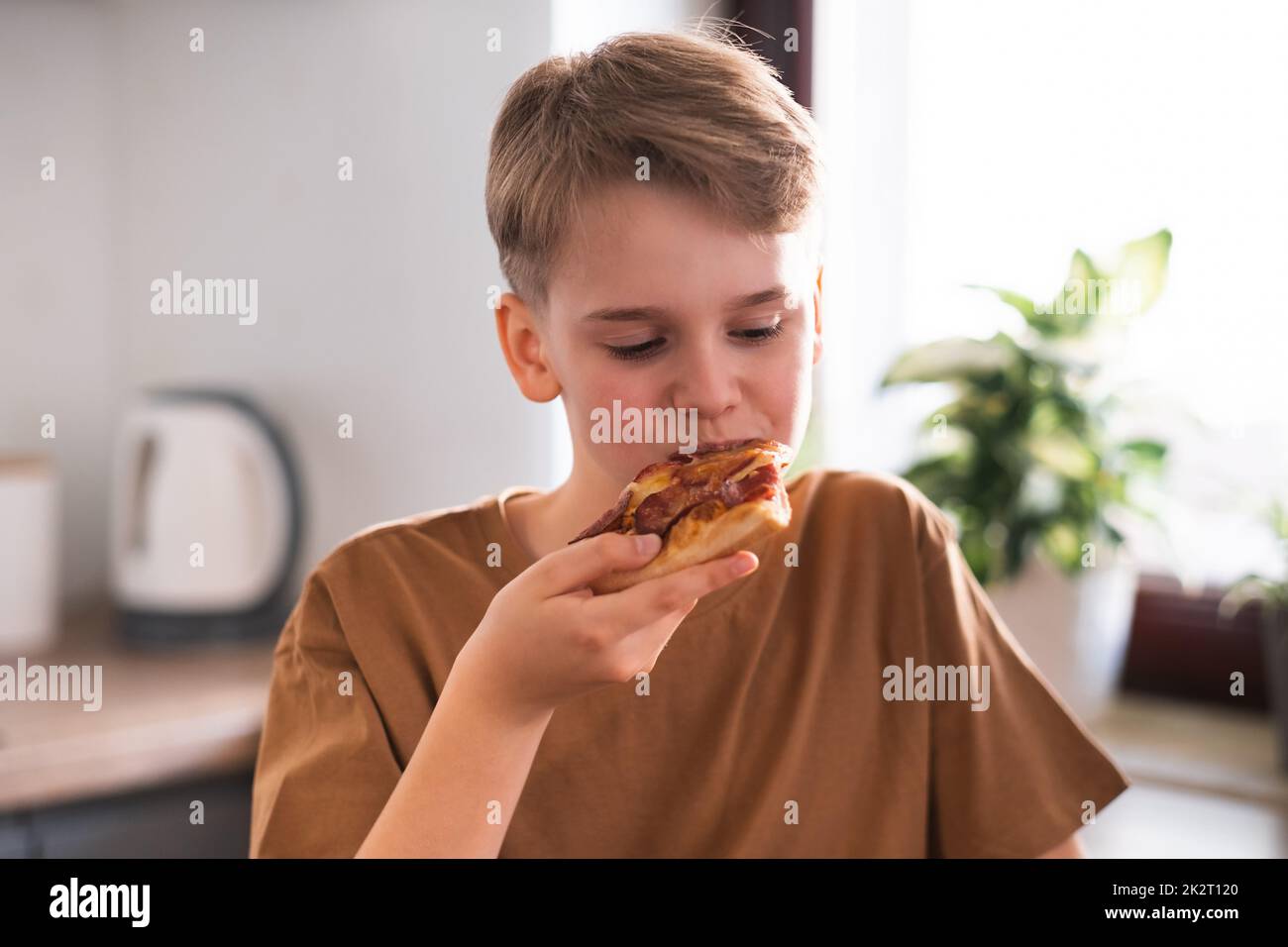 Il ragazzo con piacere mangia una deliziosa pizza in cucina a casa. Foto Stock
