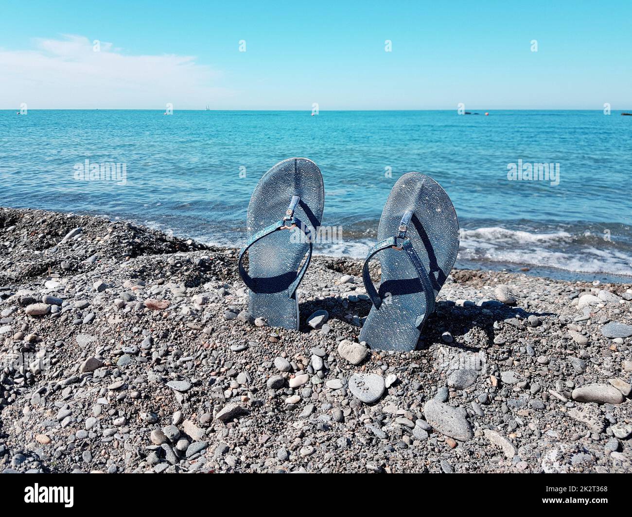 Scarpe da spiaggia blu da donna, in piedi nei ciottoli sullo sfondo del mare e del cielo blu. Primo piano, un luogo di testo, il concetto di una vacanza al mare Foto Stock