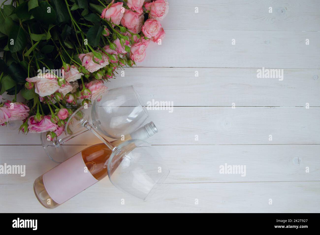 Un grande bouquet di rose rosa e una bottiglia di vino rosato con bicchieri giacciono sul lato su uno sfondo bianco di legno con un posto per il testo Foto Stock