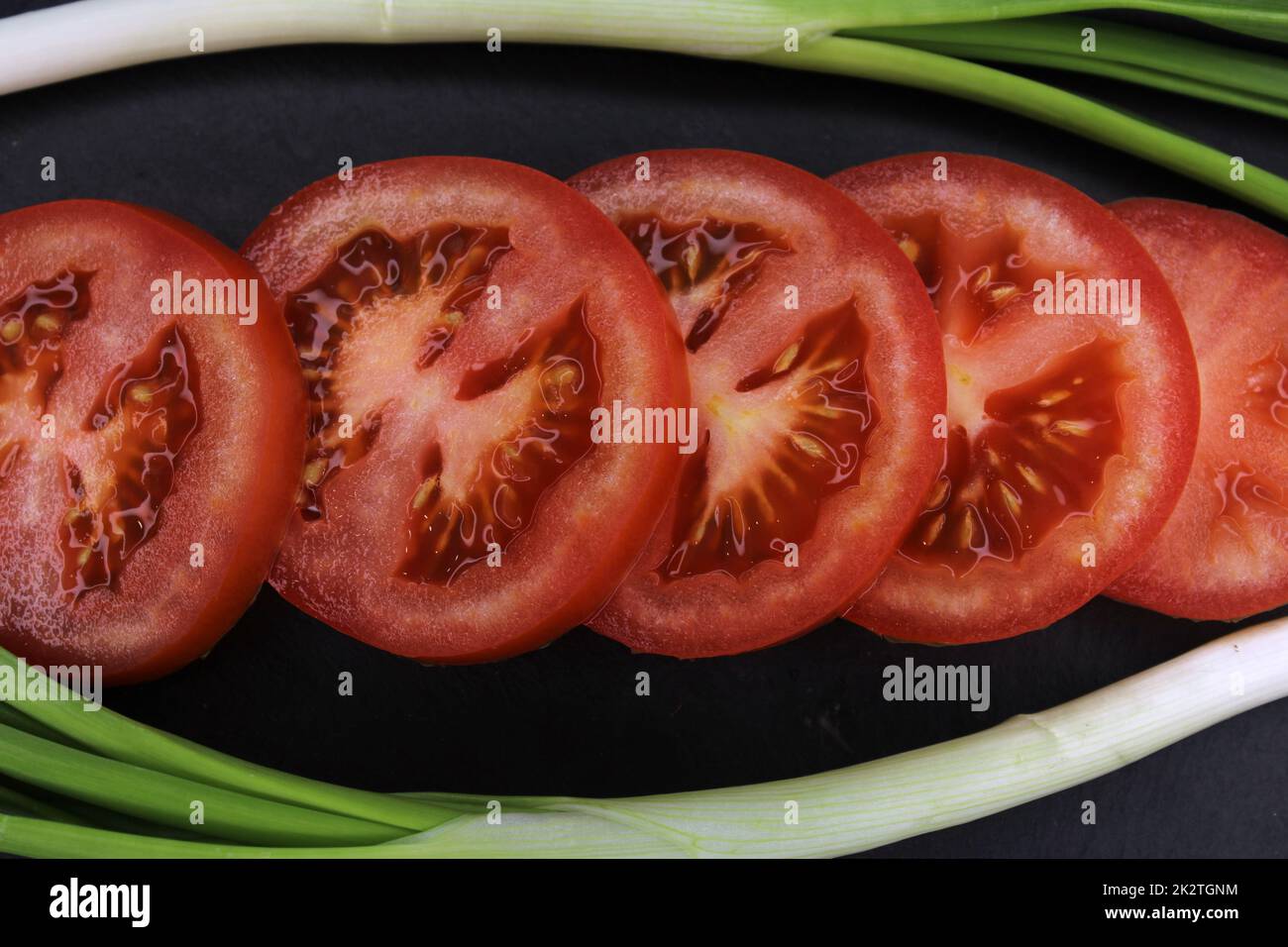 Fette di pomodoro in una cornice di cipolle verdi primo piano su sfondo nero. Il concetto di alimentazione sana. Foto Stock