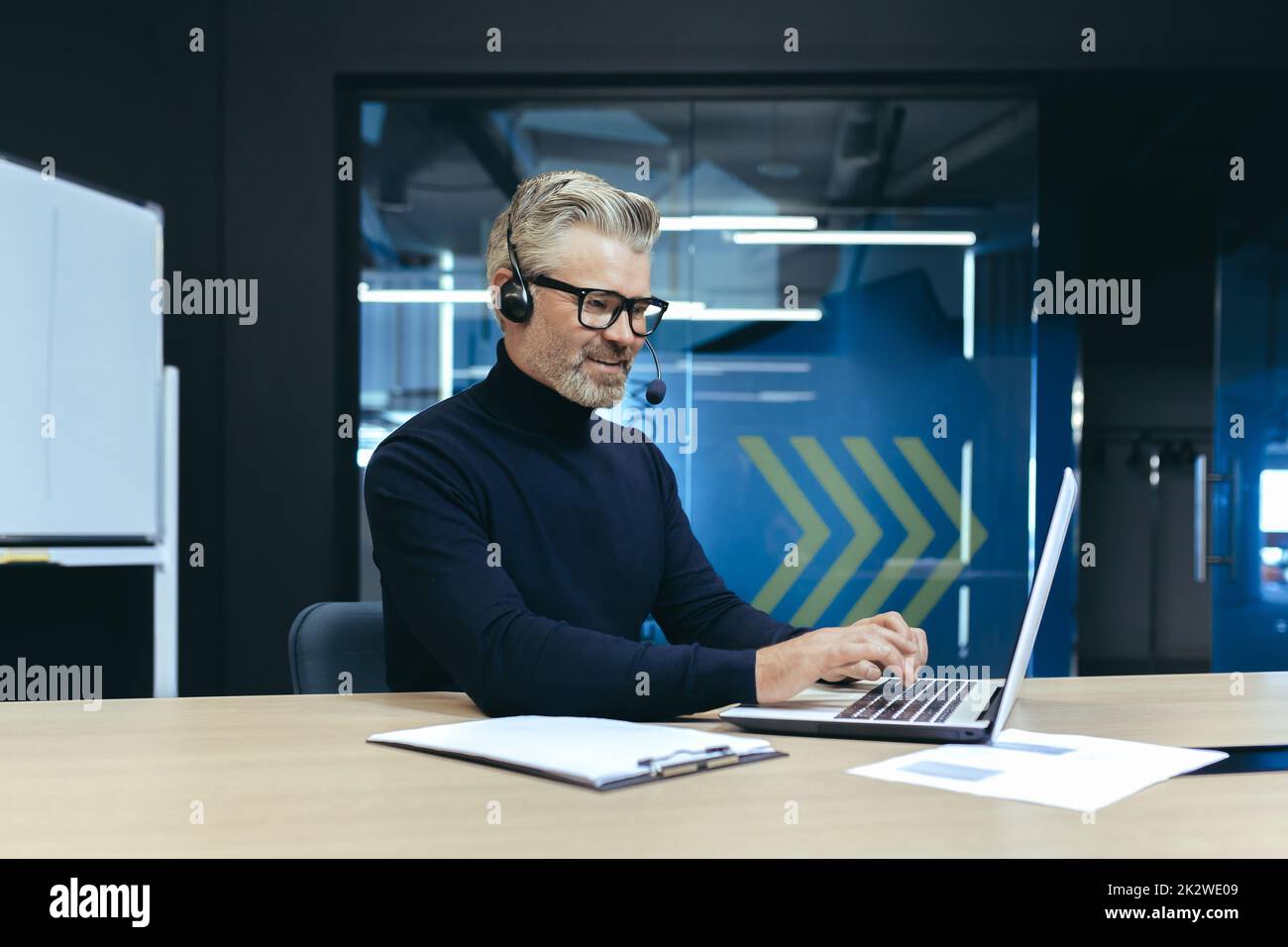 Investitore senior dai capelli grigi, uomo d'affari che lavora all'interno di un ufficio moderno utilizzando laptop e cuffie per videochiamate e riunioni a distanza con colleghi e partner. Foto Stock