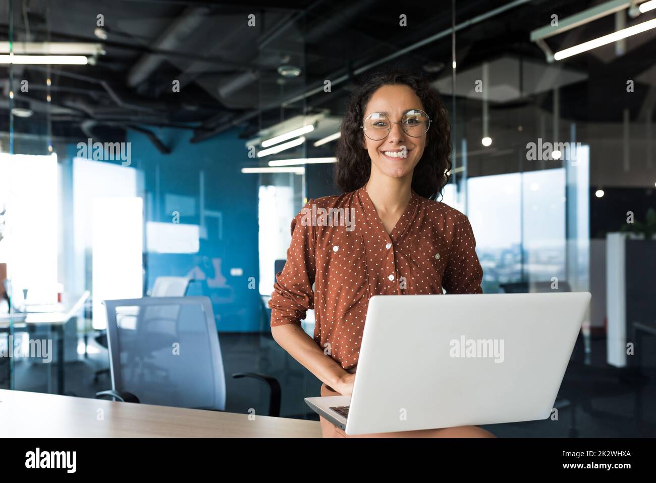 Ritratto di giovane bella donna ispanica con notebook, donna d'affari all'interno di un moderno edificio ufficio sorridendo e guardando la macchina fotografica. Foto Stock