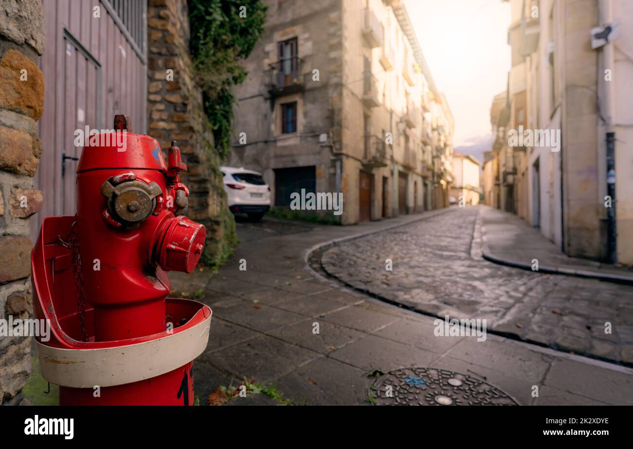 Idrante rosso fuoco sul marciapiede nella città di Onati, Spagna. Idrante antincendio su vecchio edificio sfocato, auto bianca e strada. Paesaggio urbano. Alimentazione dell'acqua per l'estintore. Sistema antincendio della città per la sicurezza. Foto Stock