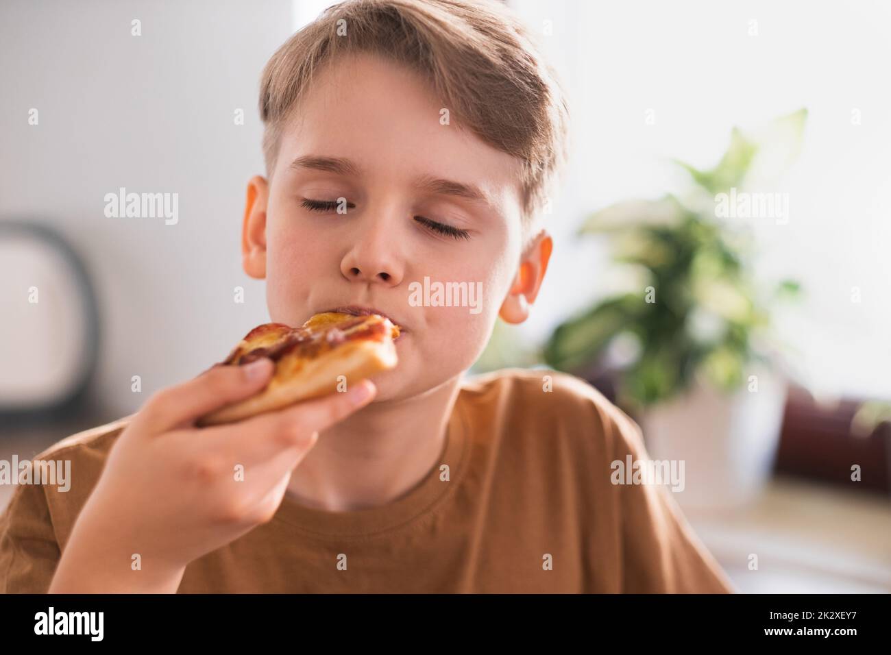 Il ragazzo con piacere mangia una deliziosa pizza in cucina a casa. Foto Stock