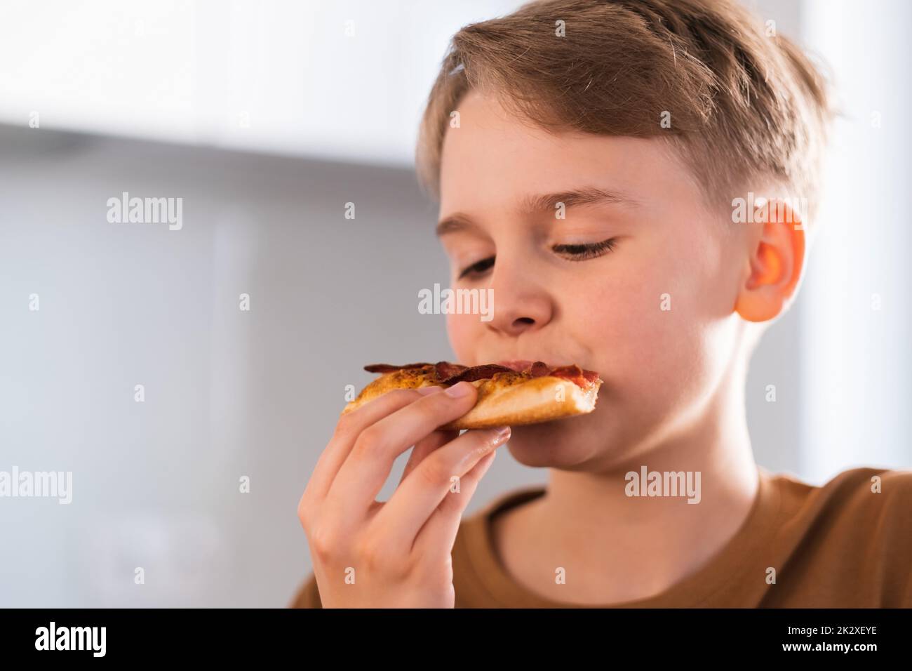 Il ragazzo con piacere mangia una deliziosa pizza in cucina a casa. Foto Stock