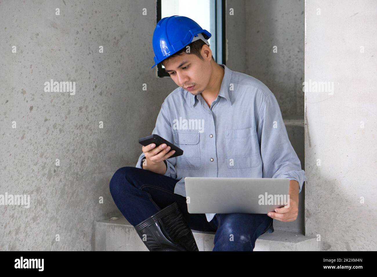 Giovane ingegnere asiatico in hardhat trasferire i dati dal telefono cellulare al computer portatile mentre si siede su una scala. Ambiente di lavoro degli ingegneri nel cantiere di progetti abitativi. Foto Stock