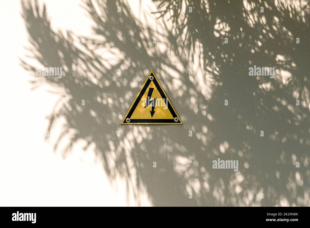 Simbolo di avvertimento a triangolo giallo ad alta tensione sulle porte in metallo grigio con ombra d'albero Foto Stock