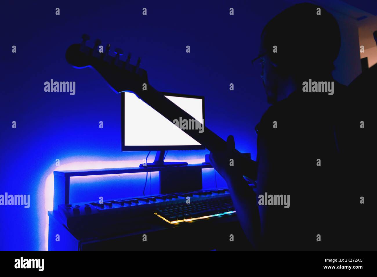 uomo al buio che lavora nel suo studio di musica di casa con luci a led blu dietro la scrivania, sta suonando la chitarra e guardando il monitor, controllando t Foto Stock