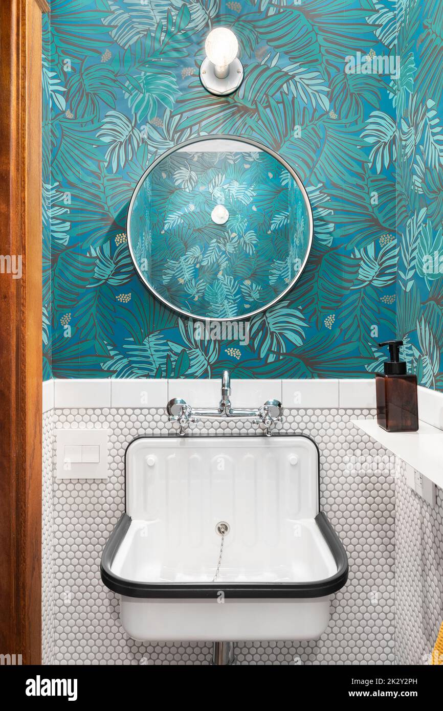 Bagno ristrutturato con carta da parati e pareti piastrellate, lavabo bianco e specchio circolare. Foto Stock