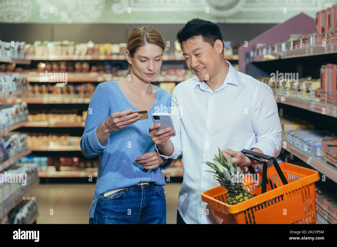 Famiglia diversa coppia asiatica uomo e donna in supermercato soddisfatto e felice shopping, utilizzando app sul telefono per selezionare i prodotti e la lista della spesa, utilizzare la carta di credito. Foto Stock