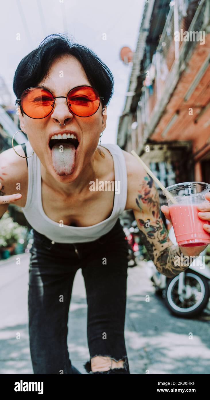Donna tatuata che esce lingua tenendo un bicchiere di bevanda Foto Stock