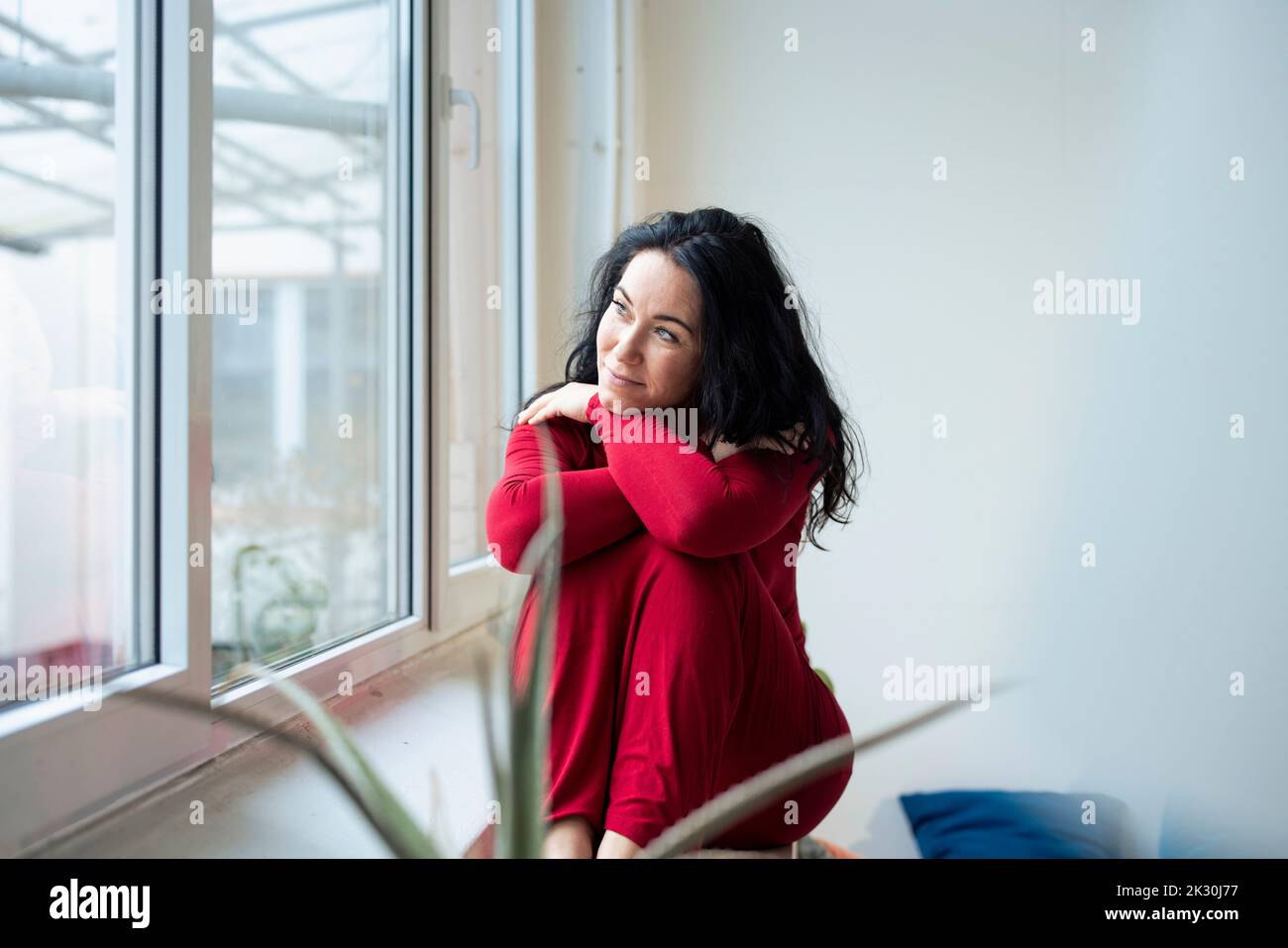 Donna premurosa che indossa un abito rosso che guarda attraverso la finestra Foto Stock