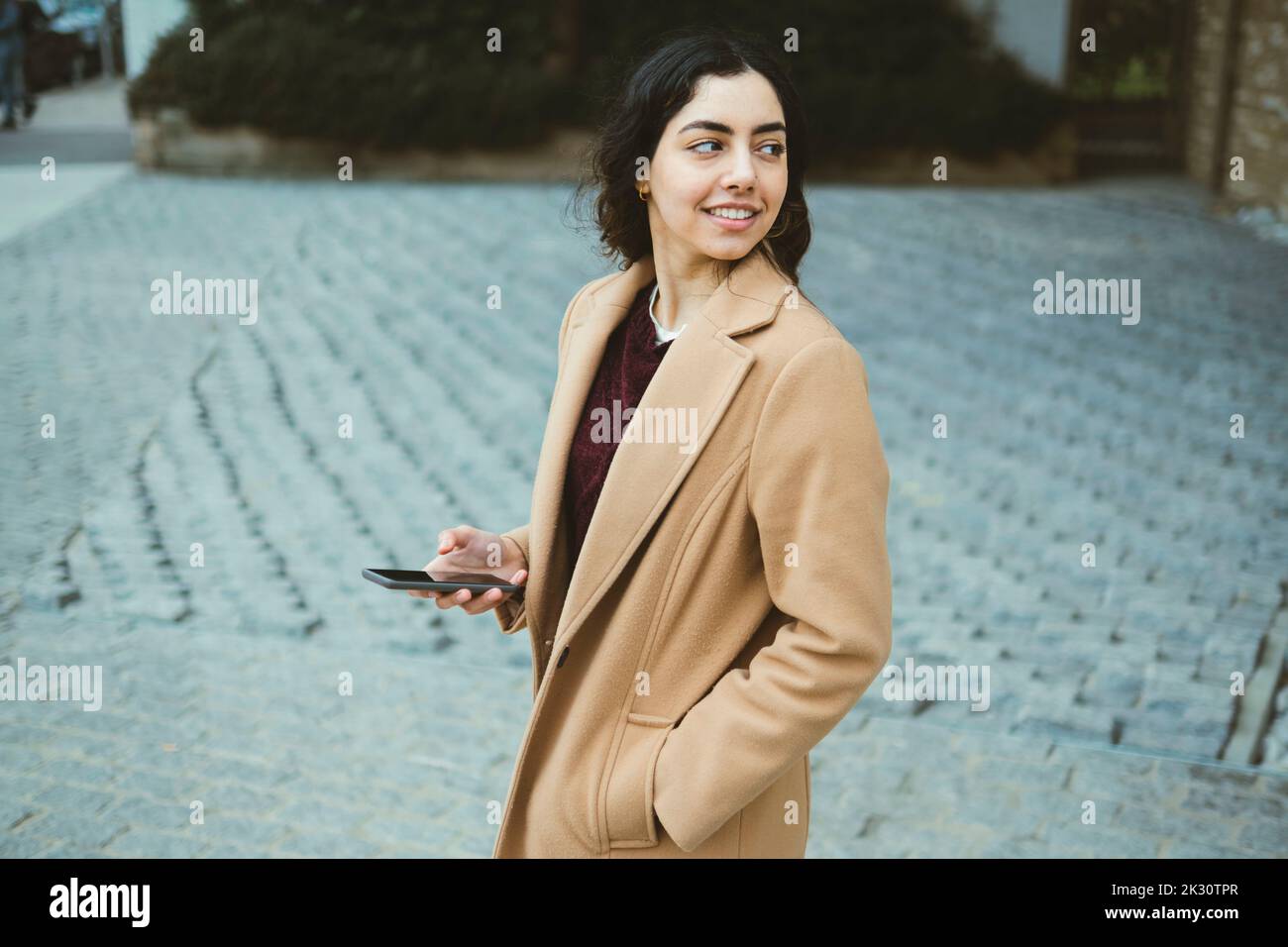 Giovane donna sorridente in trench coat con smartphone Foto Stock