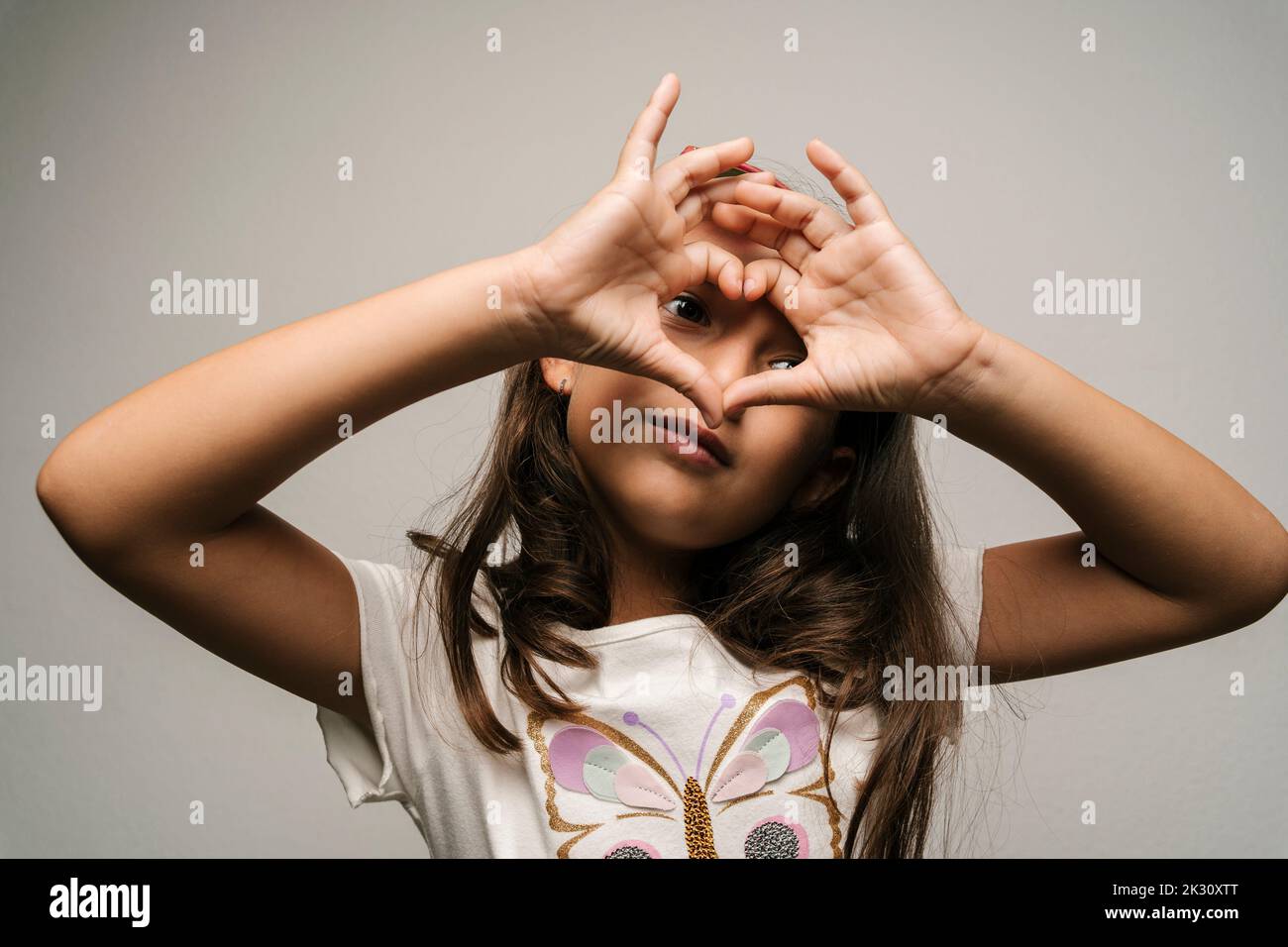 Cute ragazza gesturing forma del cuore contro sfondo grigio Foto Stock