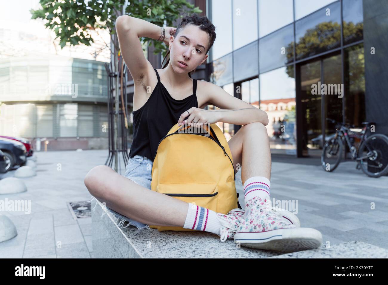 Persona non binaria con mano in capelli seduta con zaino su panca Foto Stock